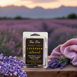 Fae Fire lavender wax melt package displayed in front of a dreamy lavender field at sunset. Black label with gold text reads 'Lavender Relaxed' against purple flowers and soft pink fabric backdrop, showcasing premium artisan crafted wax melts from Prineville, Oregon.